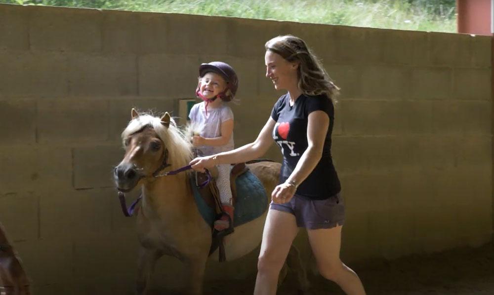 Une femme guide le poney sur lequel se trouve une jeune fille qui rigole dans le centre équestre du Couzon